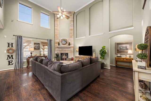 living area with dark wood-style flooring, ornamental molding, ceiling fan, a stone fireplace, and baseboards