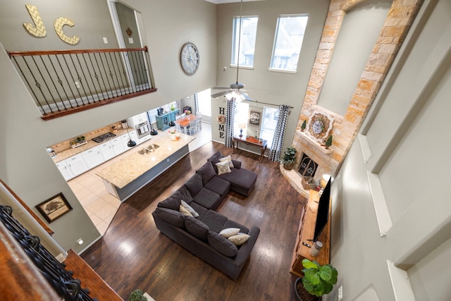 living room featuring a towering ceiling, a ceiling fan, wood finished floors, and a stone fireplace