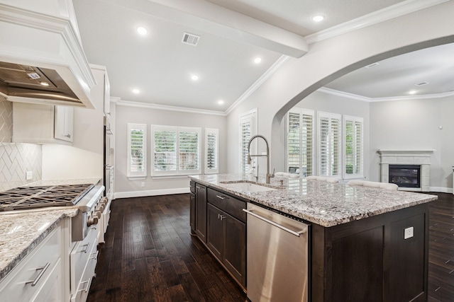 kitchen with a center island with sink, arched walkways, custom exhaust hood, stainless steel appliances, and a sink