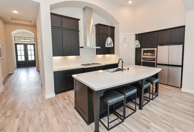 kitchen with light wood finished floors, backsplash, stainless steel appliances, wall chimney exhaust hood, and a sink