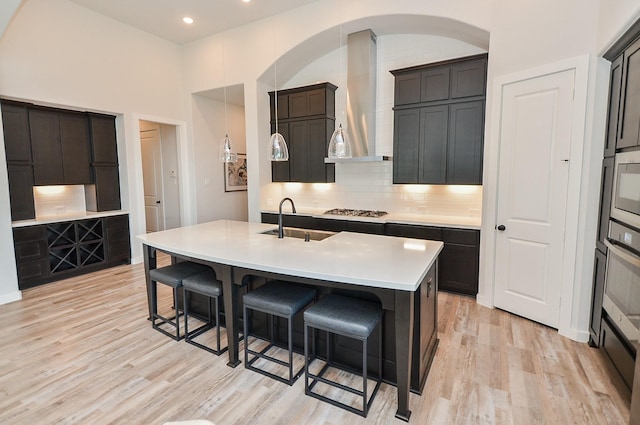 kitchen with a center island with sink, light wood-style flooring, a sink, a kitchen bar, and wall chimney range hood