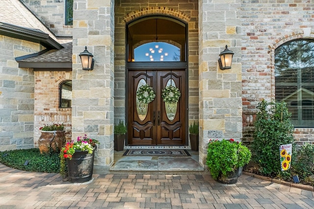 view of exterior entry with stone siding, french doors, roof with shingles, and brick siding