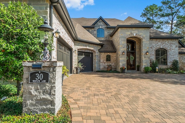 french country inspired facade with decorative driveway, roof with shingles, brick siding, an attached garage, and stone siding