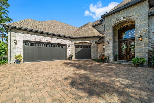 french country inspired facade with french doors, decorative driveway, roof with shingles, and an attached garage