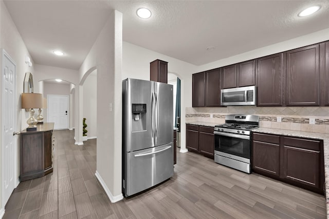 8219 Quiet Prairie Trl, Houston, TX 77049 - kitchen with stainless steel appliances, arched walkways, dark brown cabinetry, light wood-type flooring, and backsplash
