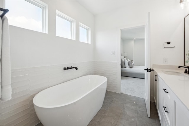 full bath with ensuite bathroom, vanity, tile walls, a freestanding bath, and wainscoting