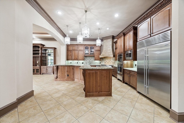 kitchen featuring built in appliances, light stone counters, a kitchen island, custom exhaust hood, and decorative backsplash