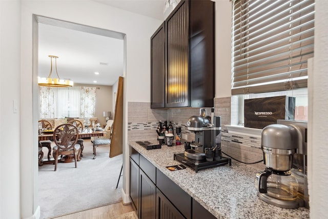 kitchen with dark brown cabinetry, light carpet, hanging light fixtures, backsplash, and light stone countertops