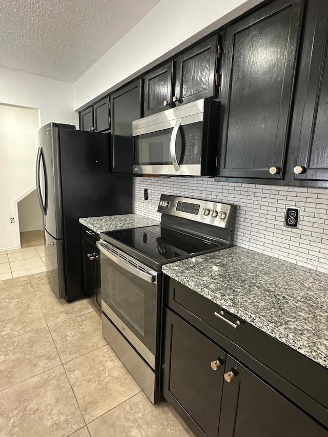 kitchen featuring light tile patterned floors, stainless steel appliances, decorative backsplash, dark cabinets, and dark stone counters