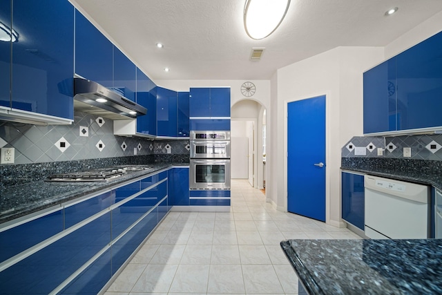 kitchen with dark stone counters, blue cabinetry, appliances with stainless steel finishes, and under cabinet range hood