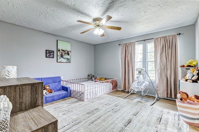 bedroom with a nursery area, a ceiling fan, light wood finished floors, and a textured ceiling