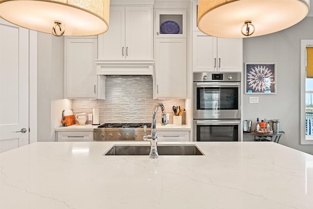 kitchen featuring double oven, a sink, white cabinets, glass insert cabinets, and custom range hood