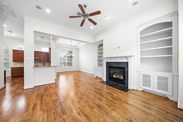 unfurnished living room with built in shelves, ceiling fan with notable chandelier, hardwood / wood-style flooring, crown molding, and a tile fireplace