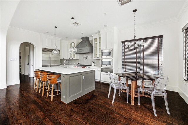 kitchen featuring custom exhaust hood, light countertops, glass insert cabinets, an island with sink, and built in appliances