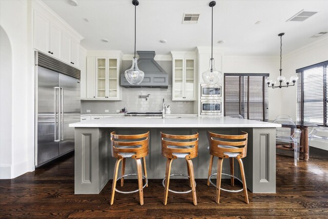 kitchen featuring a kitchen island with sink, glass insert cabinets, light countertops, and built in appliances