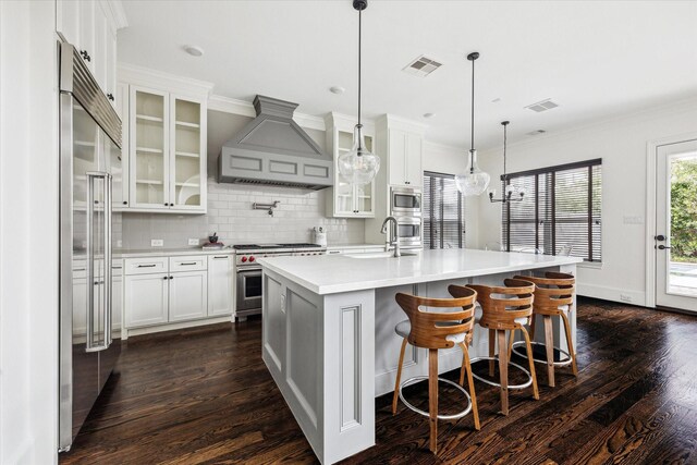 kitchen with built in appliances, a kitchen island with sink, light countertops, wall chimney range hood, and glass insert cabinets