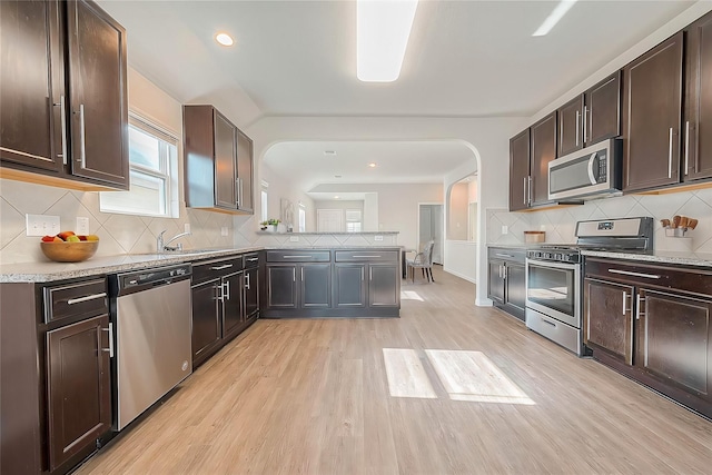 kitchen with a peninsula, light wood-style flooring, arched walkways, dark brown cabinets, and appliances with stainless steel finishes