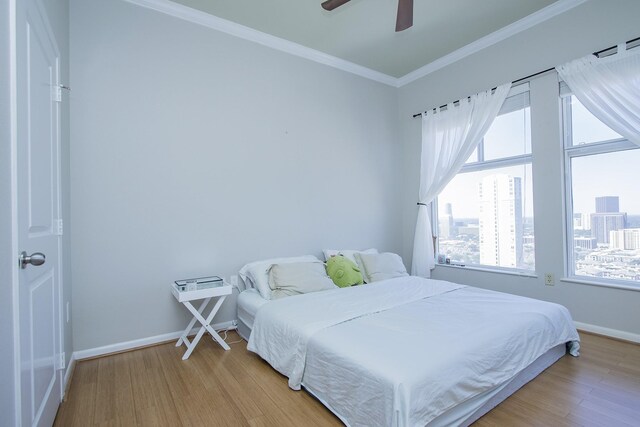 bedroom featuring a view of city, crown molding, light wood-style flooring, a ceiling fan, and baseboards