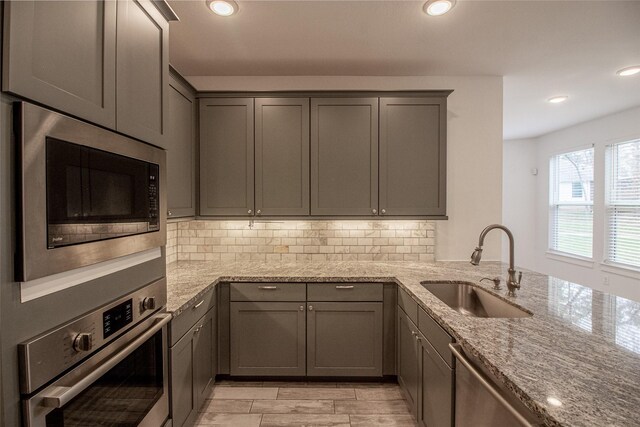 kitchen featuring gray cabinetry, a sink, appliances with stainless steel finishes, backsplash, and light stone countertops