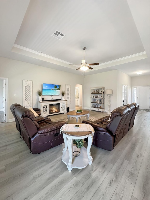 living room featuring visible vents, a raised ceiling, a ceiling fan, a glass covered fireplace, and light wood-style flooring