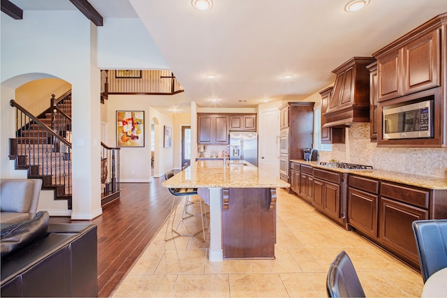 kitchen featuring a kitchen breakfast bar, built in appliances, a kitchen island with sink, premium range hood, and backsplash