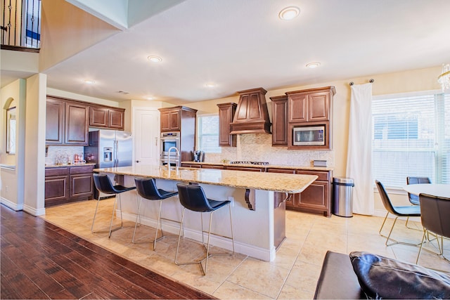 kitchen featuring an island with sink, a breakfast bar area, light stone countertops, custom exhaust hood, and stainless steel appliances