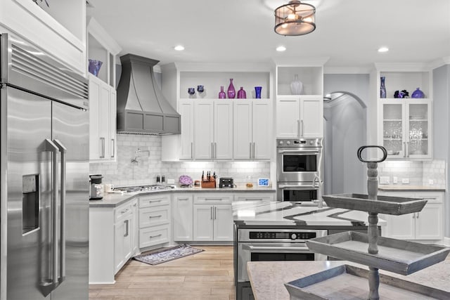 kitchen featuring stainless steel appliances, white cabinetry, backsplash, glass insert cabinets, and custom range hood