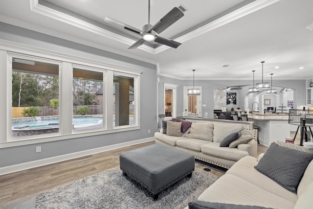 living room with a tray ceiling, crown molding, wood finished floors, baseboards, and ceiling fan with notable chandelier