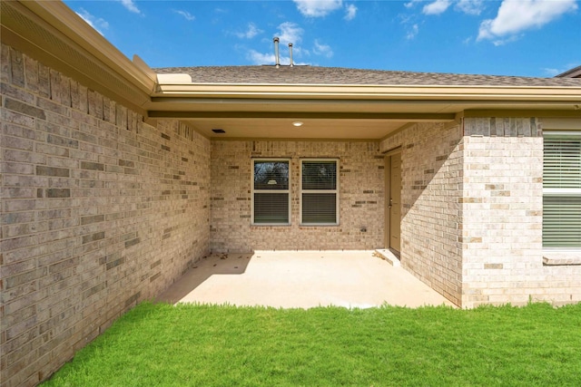 doorway to property featuring a patio area, brick siding, a yard, and roof with shingles