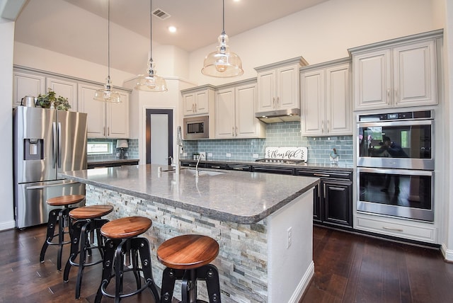 kitchen featuring under cabinet range hood, stainless steel appliances, dark wood-style flooring, visible vents, and an island with sink