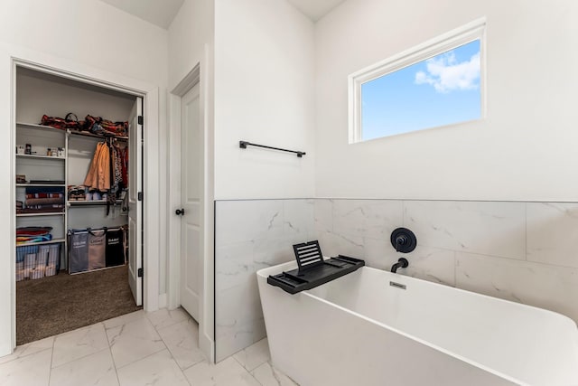 bathroom with a walk in closet, marble finish floor, tile walls, a freestanding bath, and wainscoting