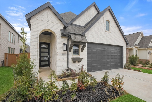 french country home with a garage, a shingled roof, concrete driveway, fence, and brick siding