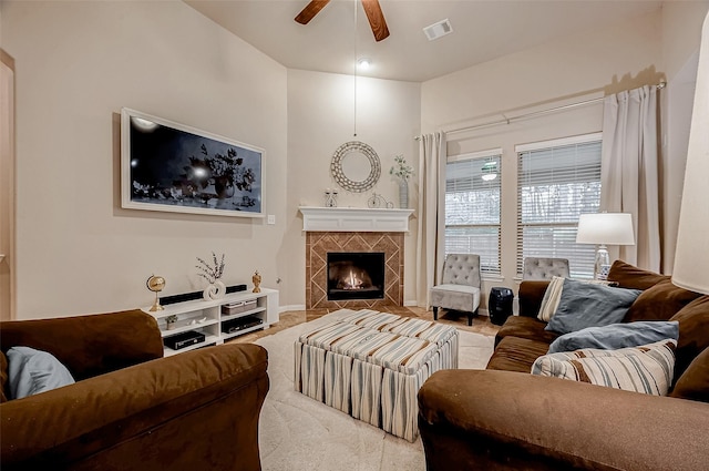 living area with ceiling fan, a tiled fireplace, visible vents, and baseboards