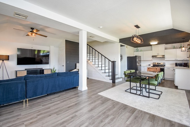 living room with visible vents, a ceiling fan, light wood-style flooring, stairs, and vaulted ceiling