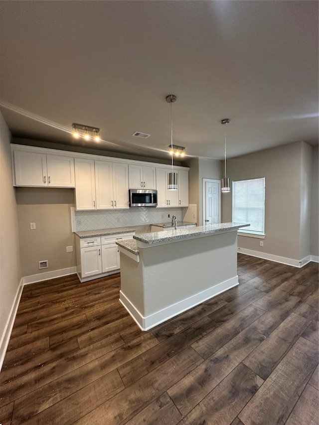 kitchen featuring light stone counters, pendant lighting, stainless steel microwave, white cabinetry, and an island with sink