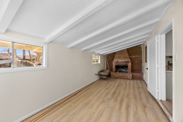 unfurnished living room with lofted ceiling with beams, a brick fireplace, a textured ceiling, light wood-type flooring, and baseboards