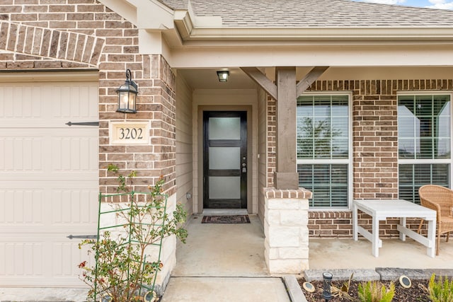 view of exterior entry with a garage, a shingled roof, and brick siding