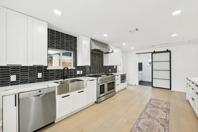 kitchen with a barn door, visible vents, stainless steel appliances, wall chimney range hood, and a sink