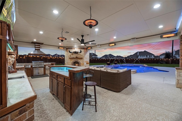 pool at dusk featuring an outdoor stone fireplace, a sink, ceiling fan, and area for grilling