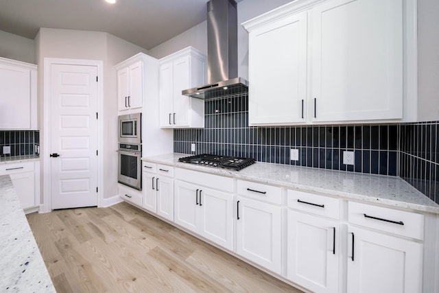 kitchen featuring light wood finished floors, appliances with stainless steel finishes, light stone counters, wall chimney range hood, and white cabinetry