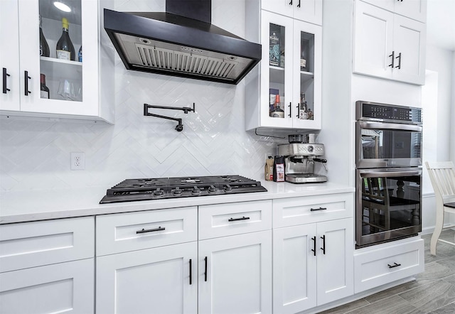 kitchen featuring light countertops, glass insert cabinets, appliances with stainless steel finishes, white cabinetry, and wall chimney range hood