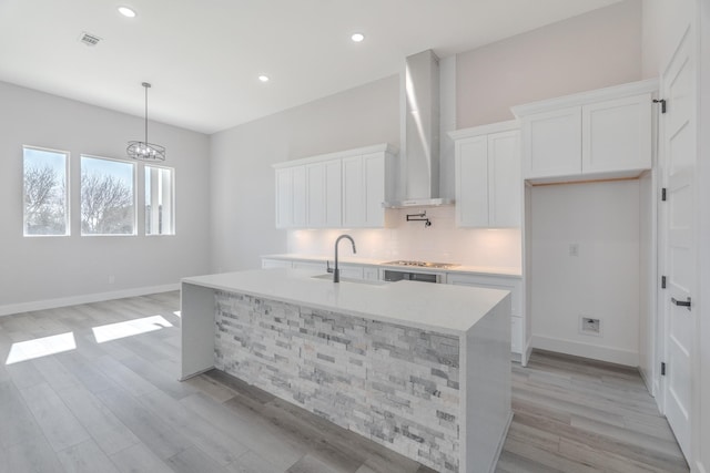 kitchen featuring light countertops, white cabinetry, a sink, wall chimney range hood, and an island with sink