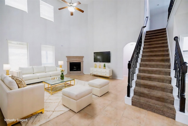 living room featuring light tile patterned floors, a fireplace with raised hearth, a ceiling fan, stairway, and a high ceiling