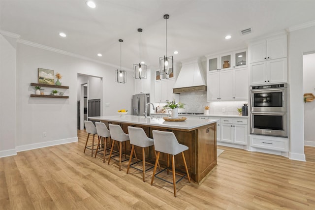 kitchen featuring custom exhaust hood, light countertops, appliances with stainless steel finishes, white cabinetry, and an island with sink