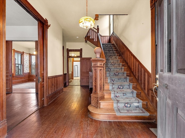 foyer entrance featuring dark wood-style flooring, a wainscoted wall, an inviting chandelier, wooden walls, and stairs