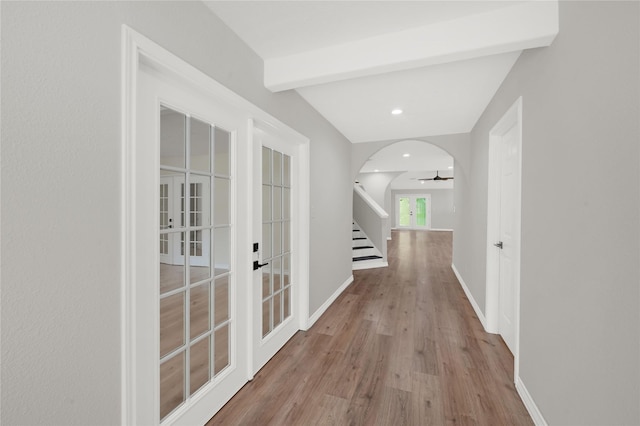 corridor featuring arched walkways, stairway, light wood-style flooring, beamed ceiling, and baseboards