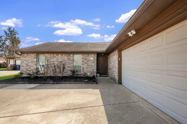 doorway to property featuring a garage, brick siding, driveway, and roof with shingles