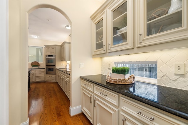 kitchen featuring arched walkways, stainless steel double oven, cream cabinetry, dark stone counters, and glass insert cabinets