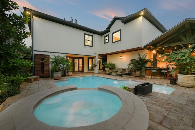 back of house at dusk with a pool with connected hot tub, a patio area, exterior bar, and stucco siding