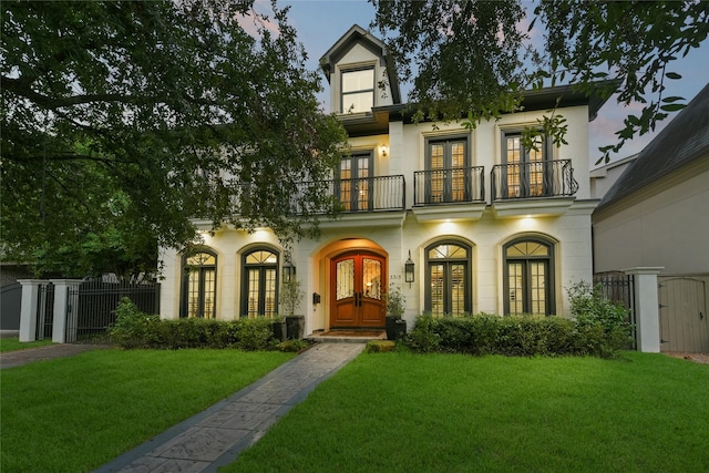 view of front of house featuring a balcony, fence, french doors, stucco siding, and a front lawn